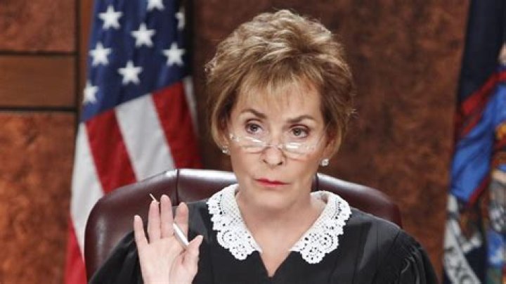 Judge Judy in court with hand raised wearing lace collar and robe