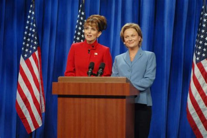 Tina Fey as Sarah Palin in a sketch from Saturday Night Live, standing at a podium with the American flag behind her.
